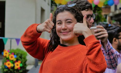 A student wearing a red jumper smiles at the camera and gives a double thumbs up